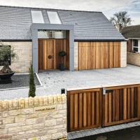 Modern house with a stone facade and wooden accents. Features a large wooden front door, a garage, and a neatly landscaped front yard with small trees and a decorative pot. A low stone wall and wooden gate enclose the property.