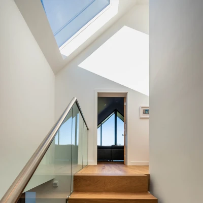 Bright, modern staircase leading to an open doorway. A large window above allows natural light to illuminate the space, while a sleek glass railing adds to the contemporary design. A hint of furniture is visible through the doorway.