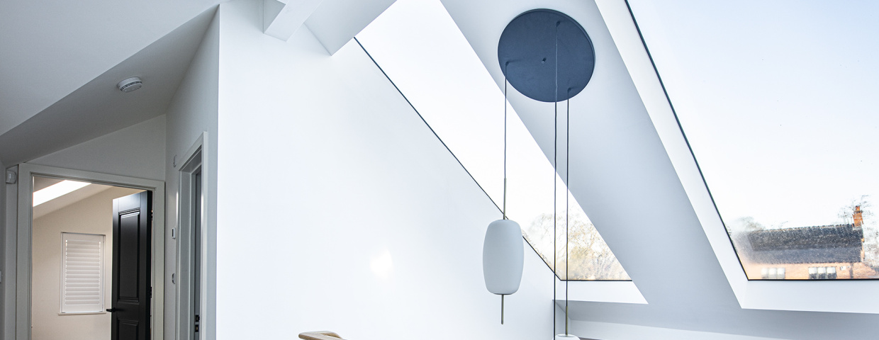 Bright interior with white walls and a high ceiling featuring large skylights. A wooden railing overlooks a lower level, with modern light fixtures hanging from the ceiling. A doorway leads to another room, and views of an outdoor area are visible through the windows.