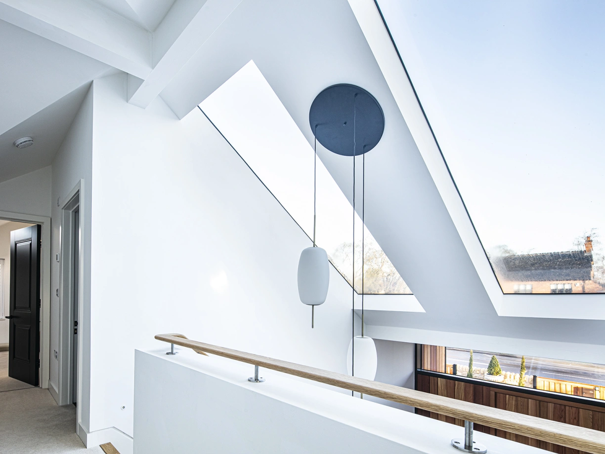 Bright, modern interior featuring a spacious hallway with a skylight and minimalist design. Light walls and a wooden railing complement the contemporary aesthetic. A circular pendant light hangs from the ceiling, illuminating the space.