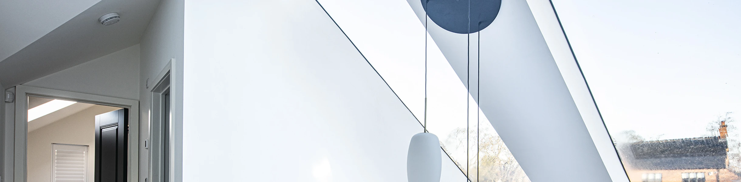 Bright interior with white walls and a high ceiling featuring large skylights. A wooden railing overlooks a lower level, with modern light fixtures hanging from the ceiling. A doorway leads to another room, and views of an outdoor area are visible through the windows.