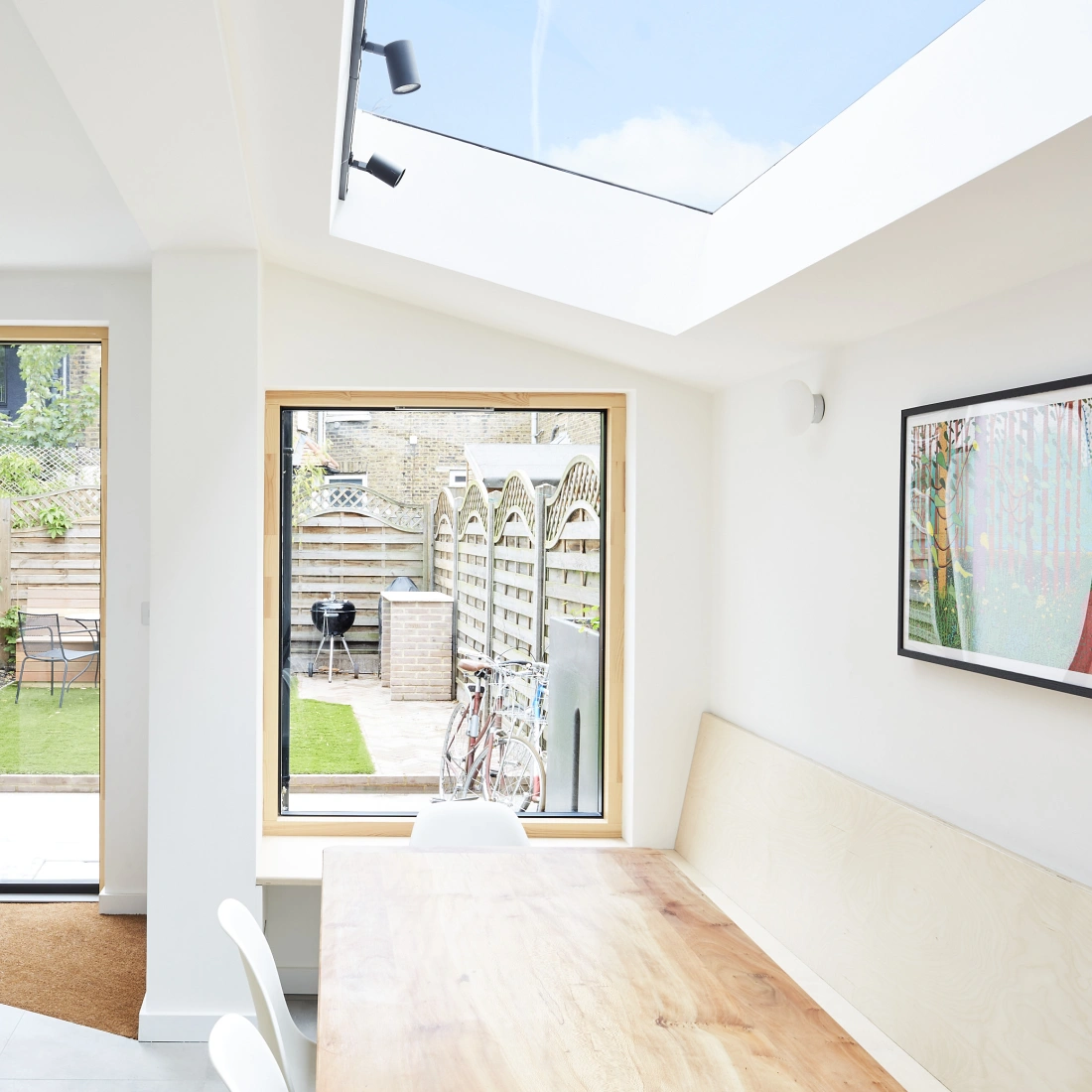 A bright and modern kitchen with a large wooden dining table, featuring a vase of flowers. The room has multiple large windows showcasing a well-maintained garden with a patio and bicycle. A skylight illuminates the space, enhancing the airy atmosphere.