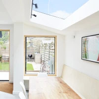 Espacio interior luminoso con una mesa de madera y sillas blancas. Ventanas grandes ofrecen vistas a un jardín con cercas de madera y un mobiliario exterior. Un techo acristalado permite la entrada de luz natural.