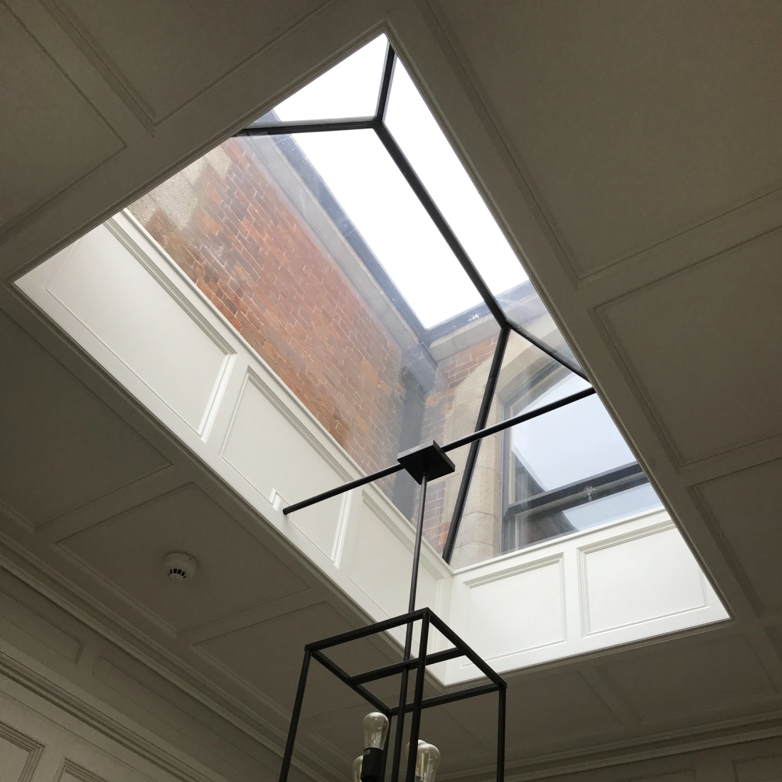 A modern square skylight allows natural light to illuminate a room with a high ceiling. Below the skylight, a geometric black chandelier with four light bulbs hangs from the ceiling. The walls are decorated with white paneling, and the exposed brick of the exterior is visible through the skylight.