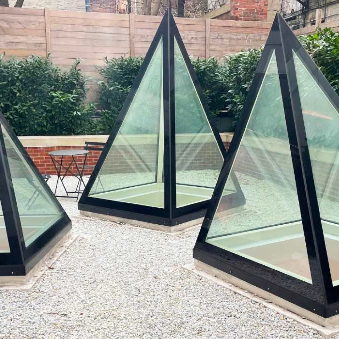 Three glass pyramids with black frames are situated in a gravel area surrounded by lush greenery and a wooden fence. A small black table and chairs are visible nearby.