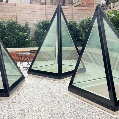 Three pyramid-shaped glass structures sit on a gravel surface, surrounded by low greenery and a brick wall. A small black table is visible nearby.