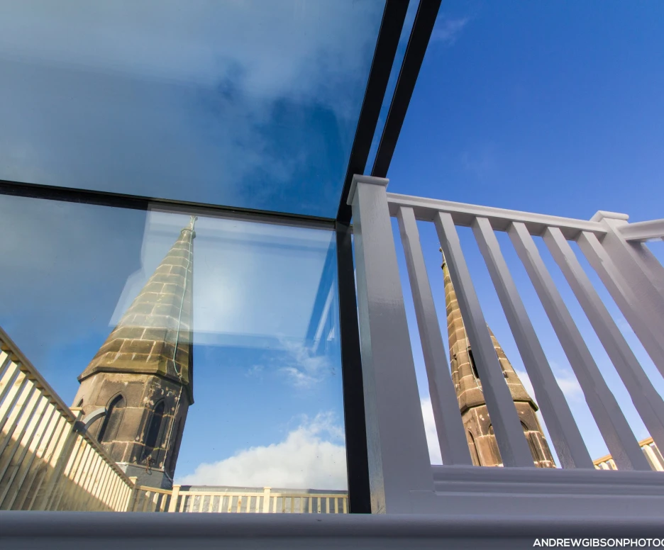 A glass structure reflects a tall church spire against a blue sky, with a white railing in the foreground creating a modern contrast to the historic architecture.
