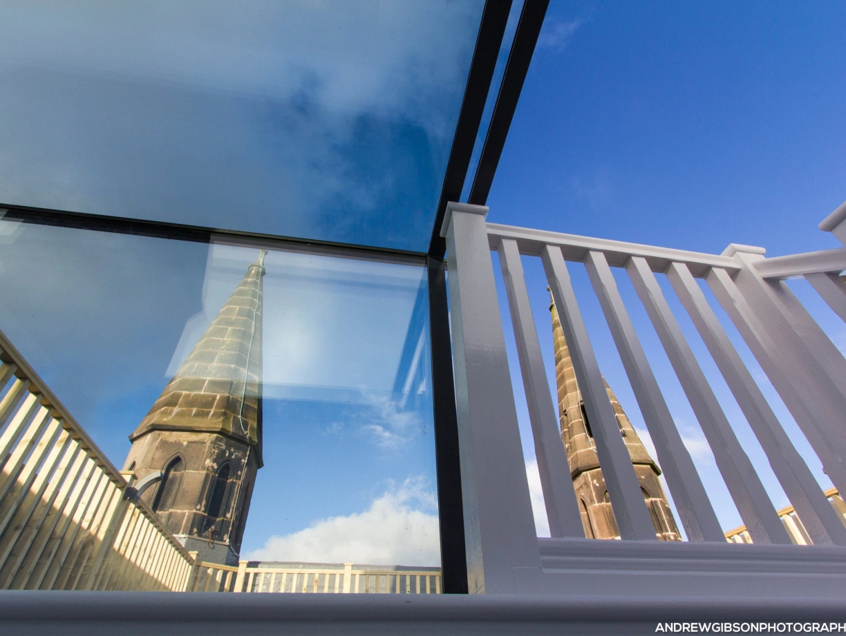 Reflection of a church tower in glass panels, framed by a white railing and a bright blue sky.