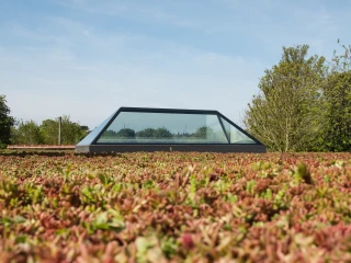 Estructura moderna con techo de cristal, rodeada de un jardín de plantas bajas y verde. El cielo es azul claro con algunas nubes.