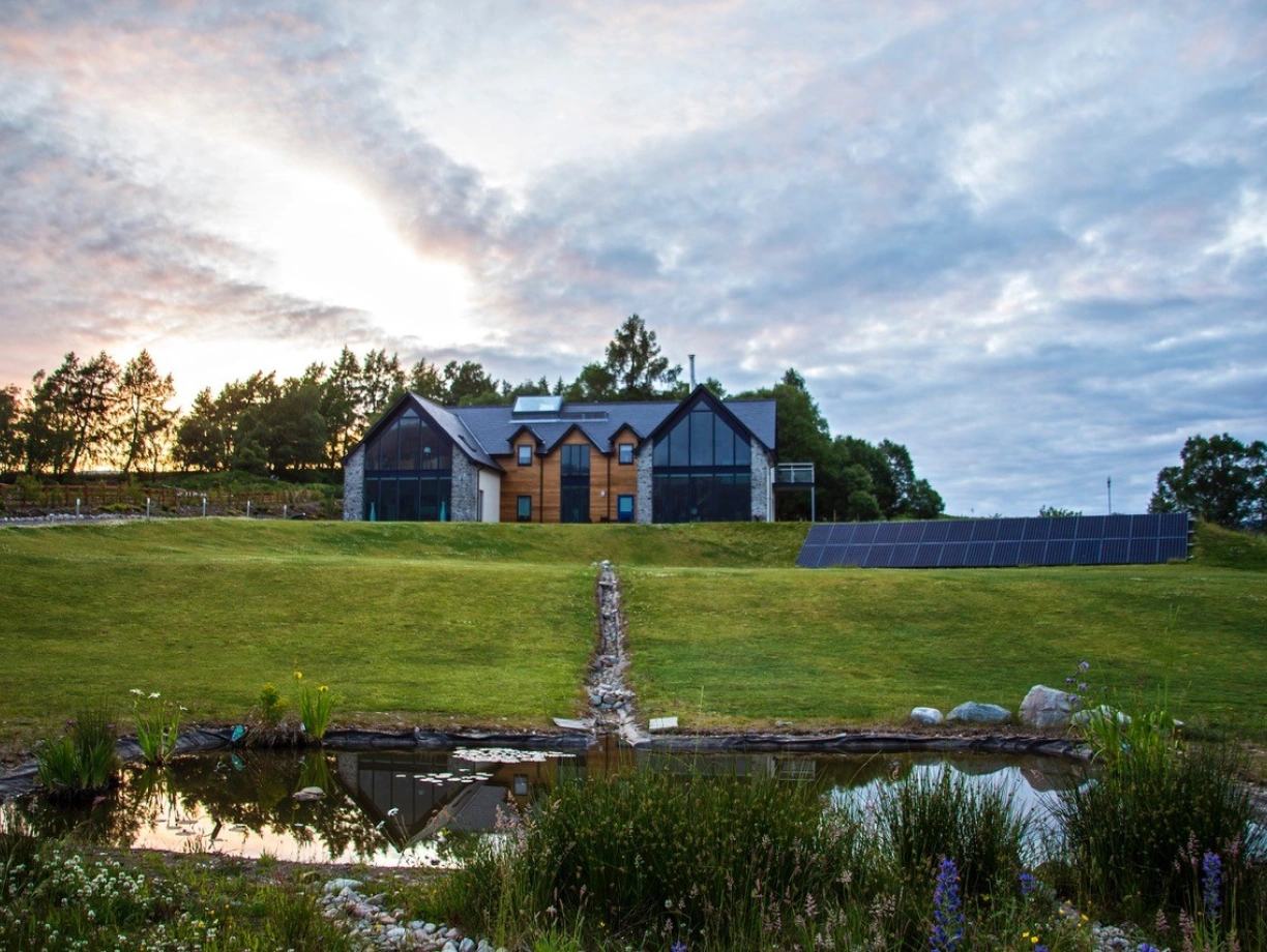 A modern house with large windows and a blend of stone and wood materials is situated on a grassy hillside. Solar panels are installed nearby, and a small pond with surrounding plants is visible in the foreground. The sky is partly cloudy, with a soft glow from sunset.