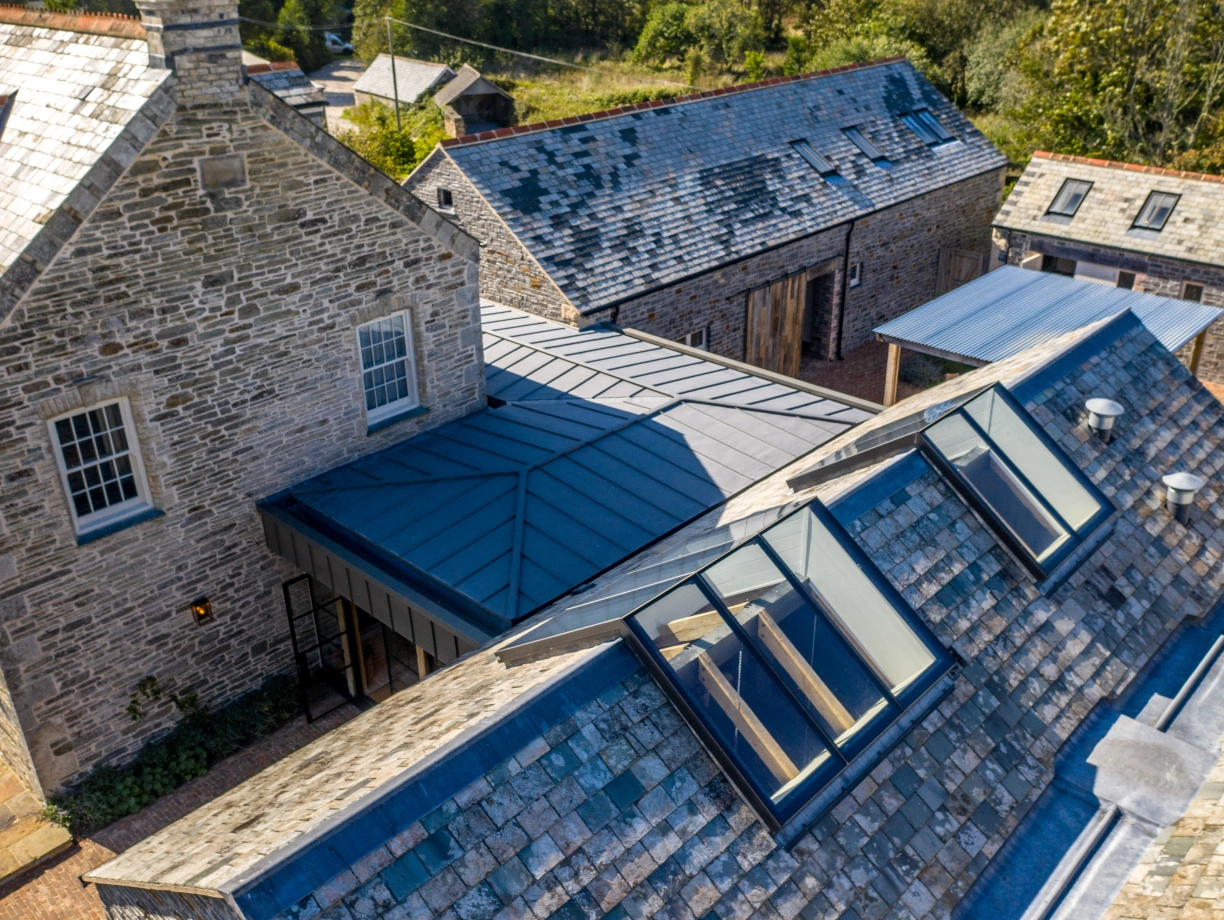 Vue aérienne d'un complexe architectural avec plusieurs bâtiments en pierre et un toit en ardoise. L'un des toits présente des lucarnes modernes, tandis que des entrepôts en bois se trouvent en arrière-plan. La verdure environnante ajoute une touche naturelle.