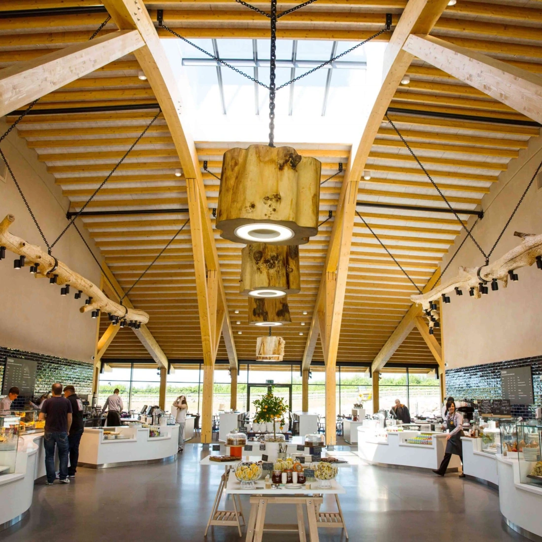 Interior de un restaurante moderno con techos altos y vigas de madera. Hay varias estaciones de comida y personas sirviéndose. La iluminación es brillante y hay grandes ventanales que permiten la entrada de luz natural.