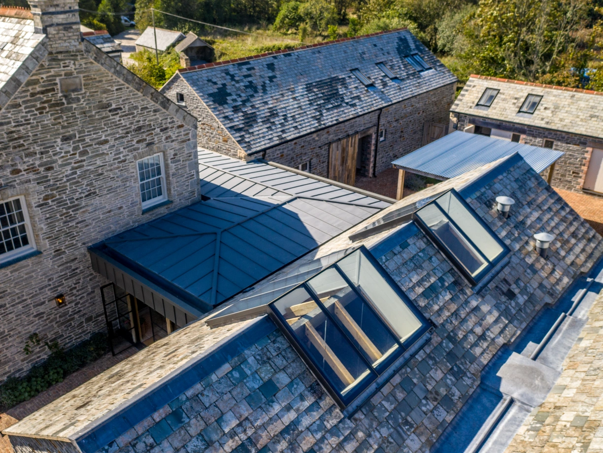 Aerial view of a rustic stone building with a slate roof, surrounded by other structures with wooden facades. Sunlight illuminates the scene, highlighting the contrasting materials and textures.