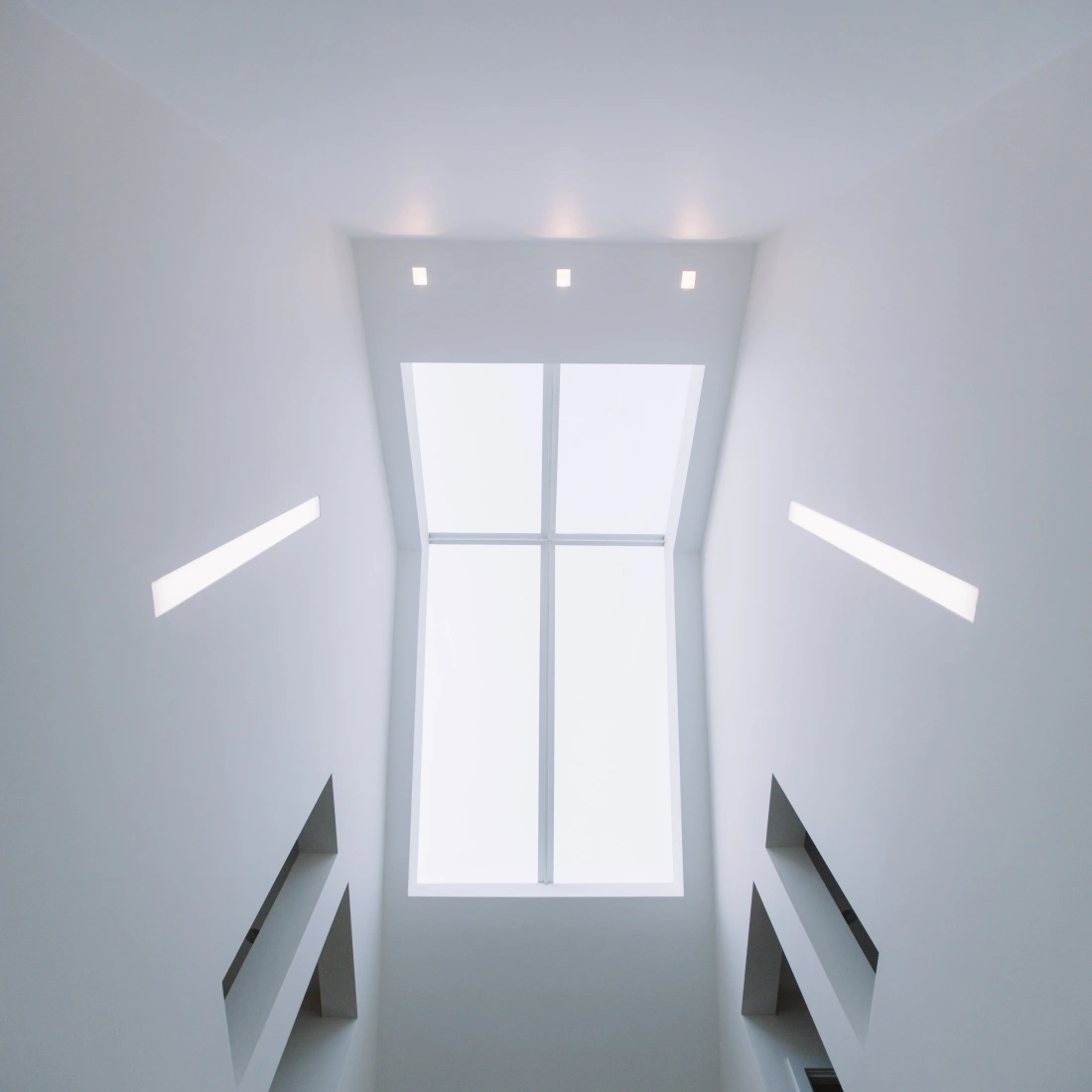 Blick nach oben in einen modern gestalteten Raum mit weißen Wänden und dreieckigen Fenstern, die Tageslicht hereinlassen. An der Decke befinden sich mehrere gleichmäßig verteilte Lichtquellen.