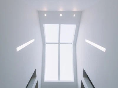 A view looking upward at a bright, minimalist stairwell. The ceiling is mostly white, featuring recessed lighting and a large rectangular window that lets in natural light.