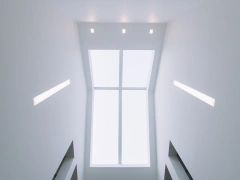 A view looking up at a bright skylight surrounded by minimalist white walls, with recessed lights illuminating the space.