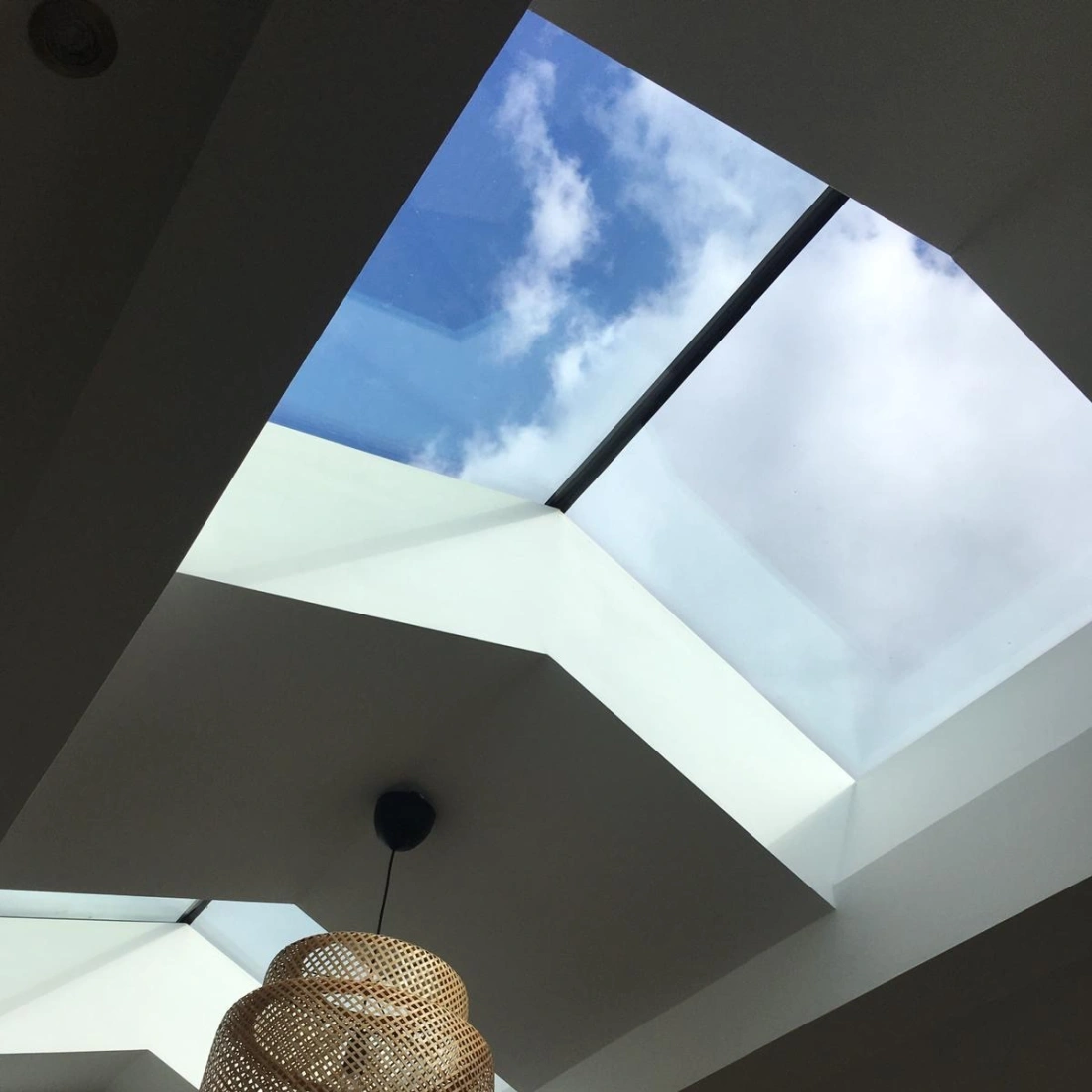 Ein modernes Dachfenster zeigt den blauen Himmel mit einigen Wolken. Darunter hängt eine dekorative Pendelleuchte aus Rattan.