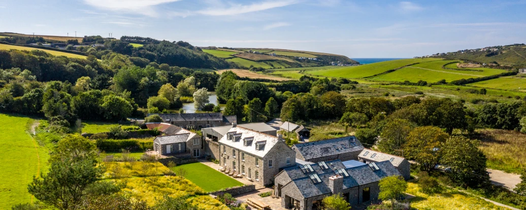 Une vue aérienne d'une grande maison en pierre entourée de verdure, avec des champs et la mer en arrière-plan. Le ciel est clair et ensoleillé.