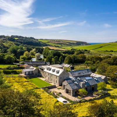 Une vue aérienne d'une grande maison en pierre entourée de verdure, avec des champs et la mer en arrière-plan. Le ciel est clair et ensoleillé.