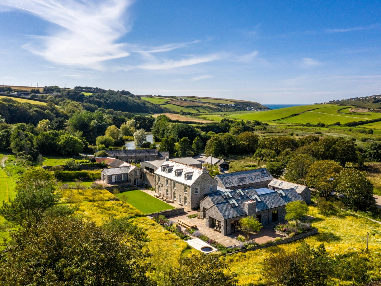 Une vue aérienne d'une grande maison en pierre entourée de verdure, avec des champs et la mer en arrière-plan. Le ciel est clair et ensoleillé.