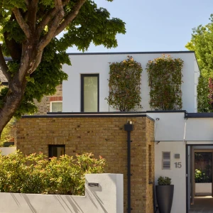 A modern two-story house featuring a combination of brick and white exterior. The upper level is adorned with greenery, while the lower level includes a front garden and a wooden gate. Tall trees and bushes surround the property, creating a lush environment.