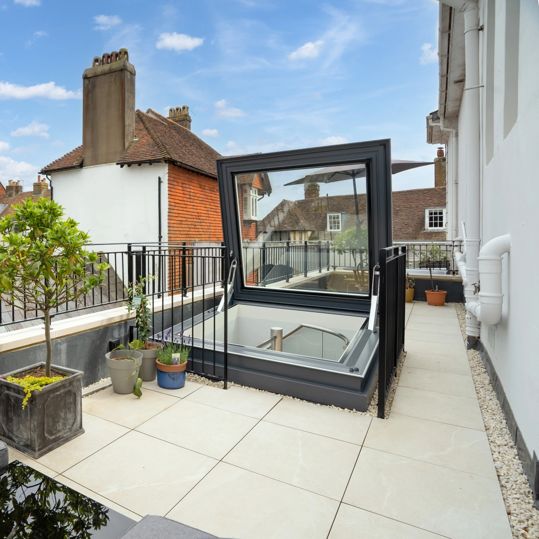 Terrasse moderne avec des carreaux en pierre, un petit arbre dans un pot et un espace lounge. Un puits de lumière est ouvert, offrant une vue sur le ciel bleu parsemé de nuages.