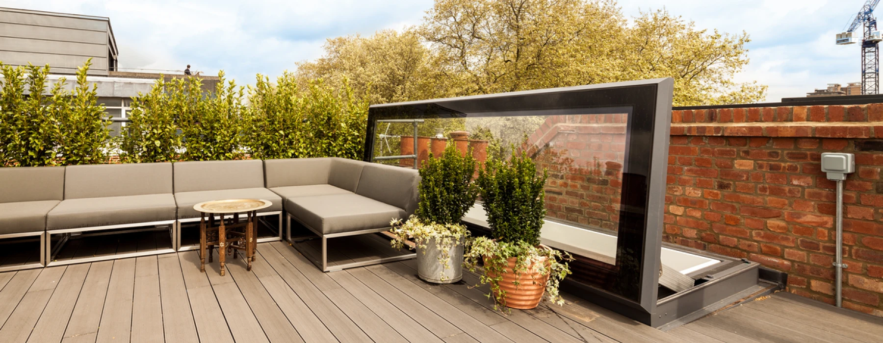 A stylish rooftop terrace featuring a modern gray sectional sofa, a small round table, and potted plants. The area is surrounded by greenery and includes a glass windbreak. Various buildings and a crane are visible in the background under a blue sky.