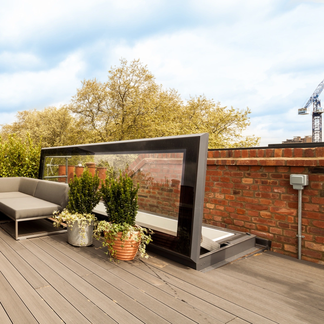 A stylish rooftop terrace featuring a modern gray sectional sofa, a small round table, and potted plants. The area is surrounded by greenery and includes a glass windbreak. Various buildings and a crane are visible in the background under a blue sky.