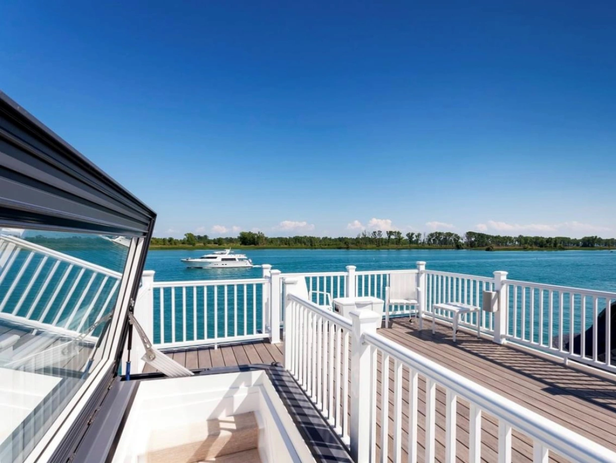 A scenic view from a dock overlooking a calm blue waterway, with a boat cruising in the distance and lush greenery lining the shore under a clear blue sky.