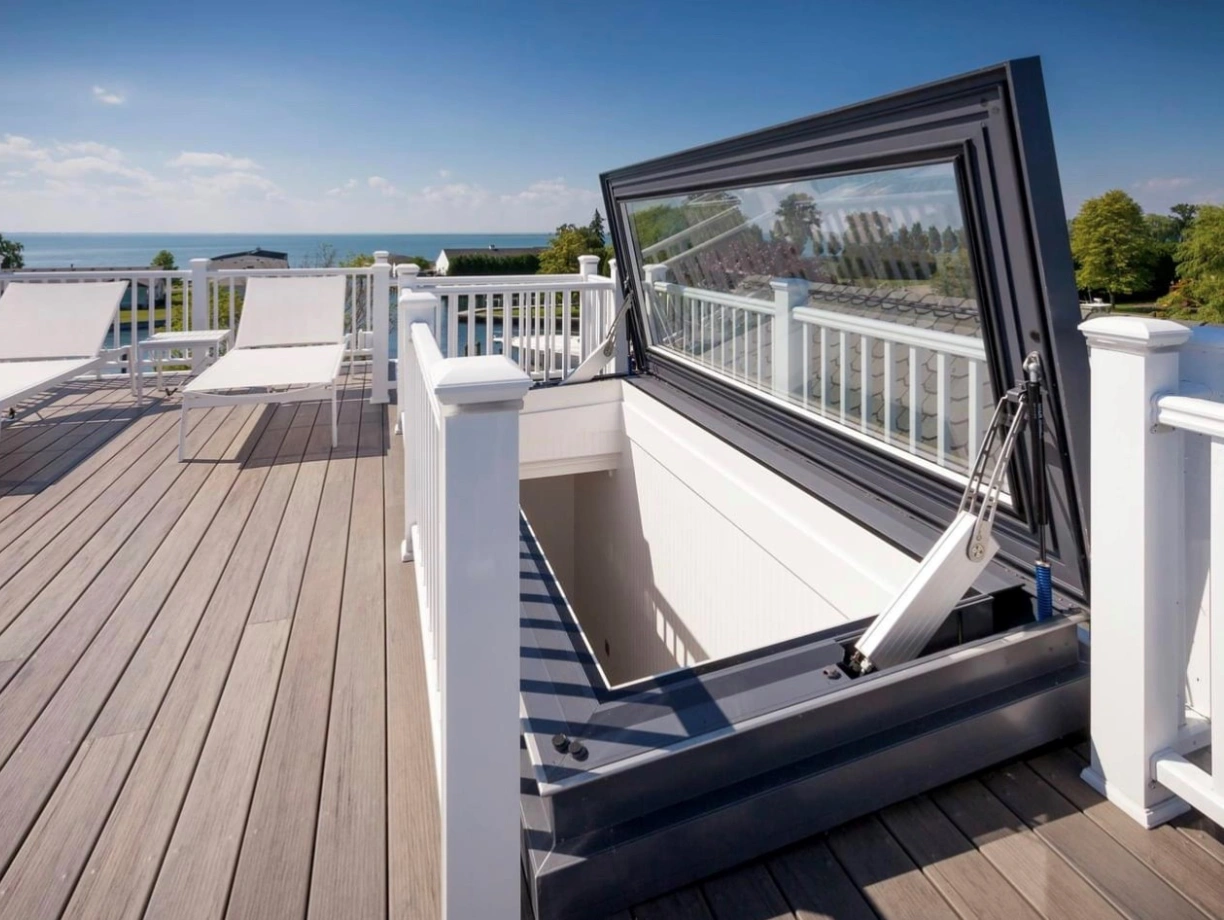 A rooftop with a large hatch opened, revealing a view of the sky and distant water. Two lounge chairs are positioned on the wooden deck, surrounded by a white railing.