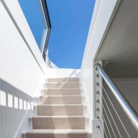 Un escalier blanc avec un tapis beige mène à un espace lumineux sous un ciel bleu. La main courante en métal ajoute une touche moderne.