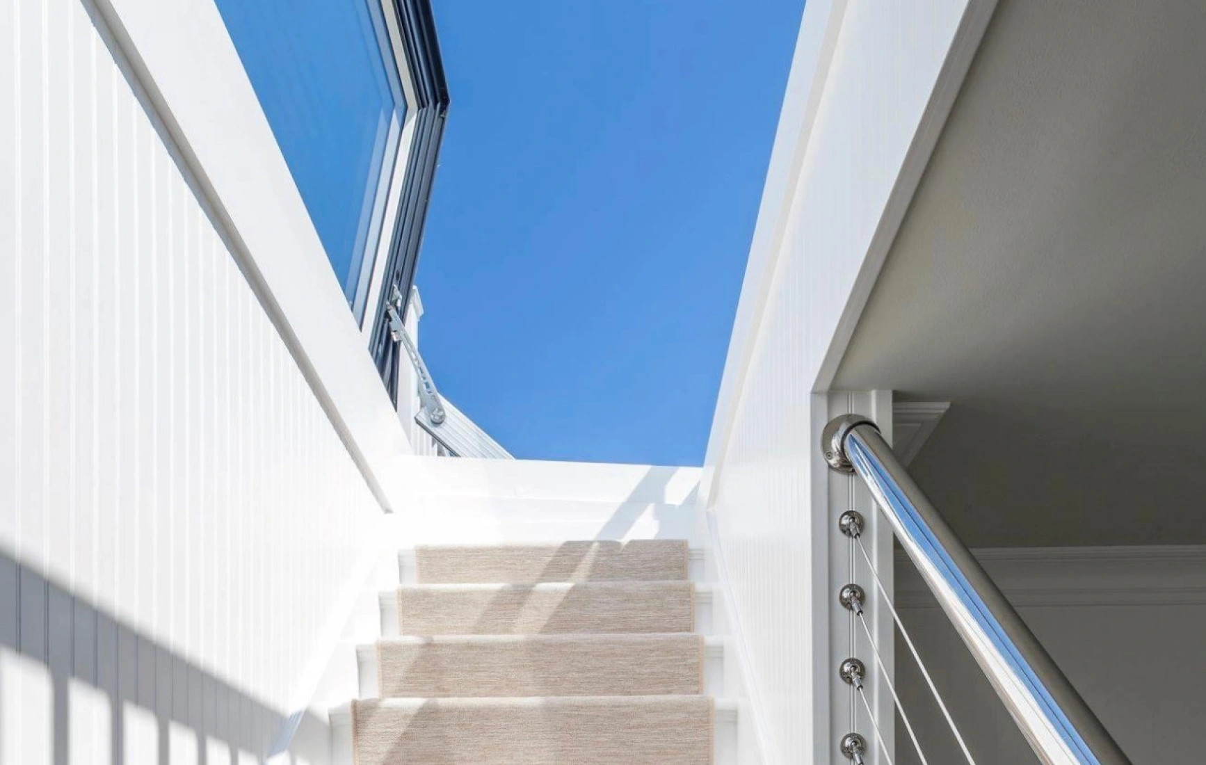 Staircase with white walls and a light-colored carpet runner leading up to a bright blue sky visible through a large window. Modern railing with stainless steel accents.