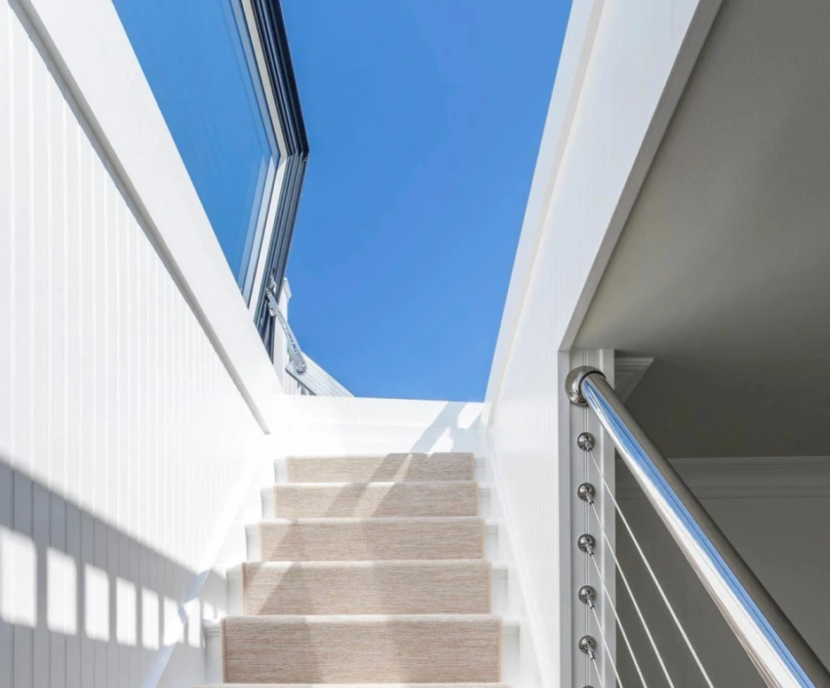 Staircase with white walls and a light-colored carpet runner leading up to a bright blue sky visible through a large window. Modern railing with stainless steel accents.
