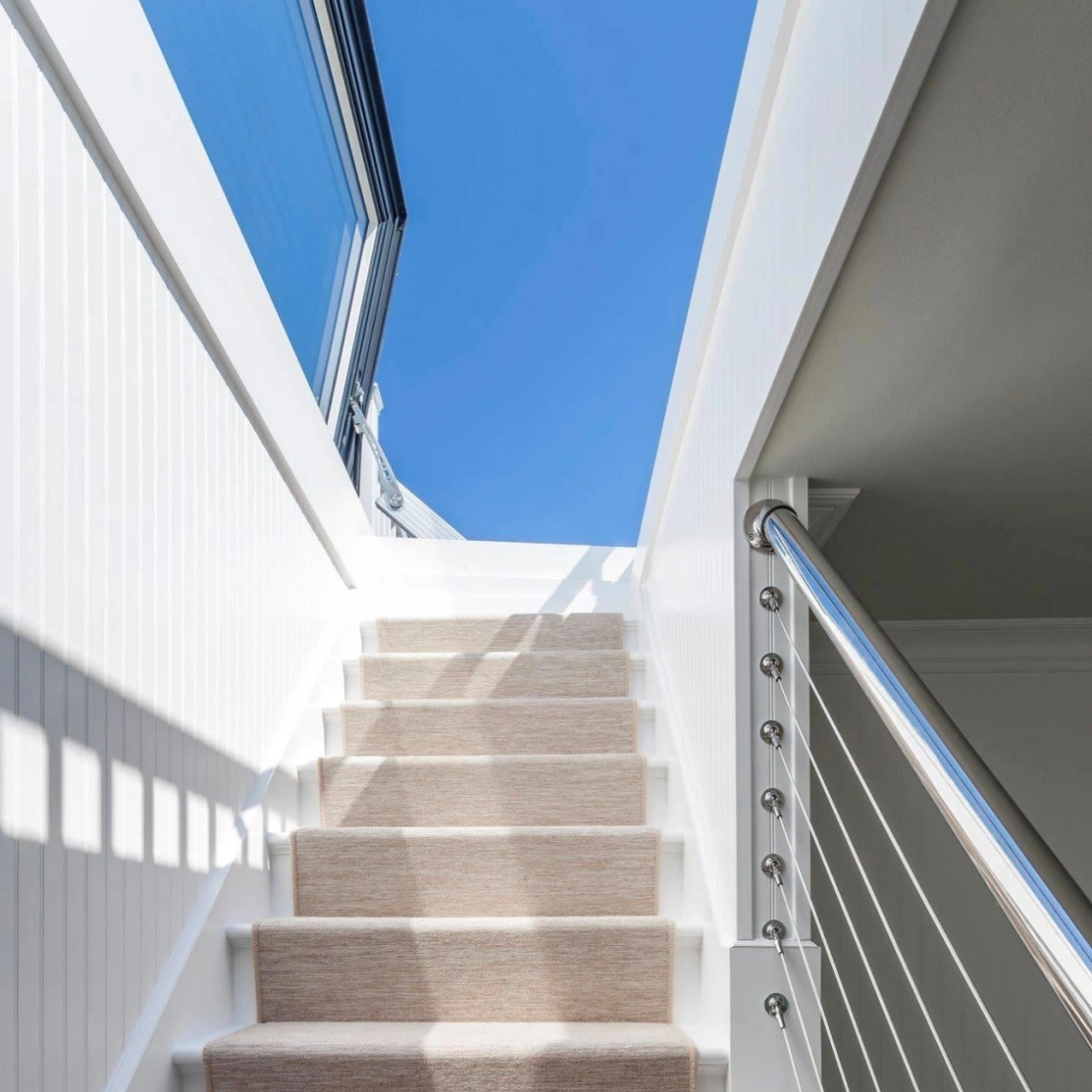 Un escalier blanc avec un tapis beige mène à un espace lumineux sous un ciel bleu. La main courante en métal ajoute une touche moderne.