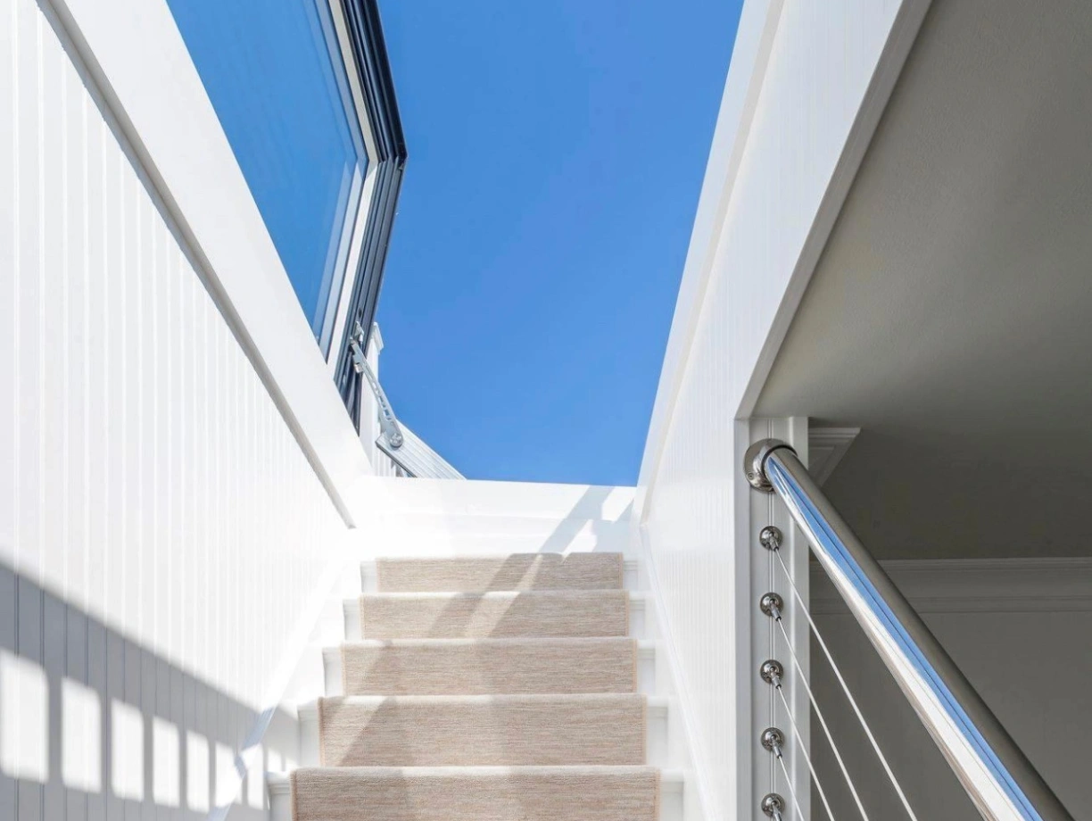 Un escalier blanc avec un tapis beige mène à un espace lumineux sous un ciel bleu. La main courante en métal ajoute une touche moderne.