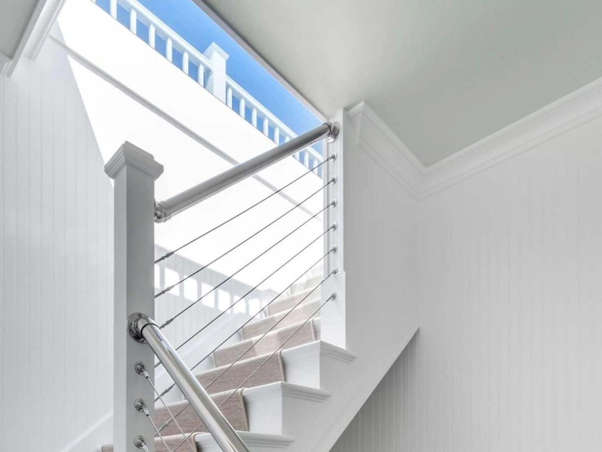 A bright, modern staircase with white walls and a stylish metal railing. Natural light filters through a window at the top, illuminating the carpeted steps. A framed picture hangs on the wall near the base of the stairs.