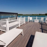 A spacious wooden deck with white lounge chairs and railings overlooks a calm blue body of water, under a clear blue sky.
