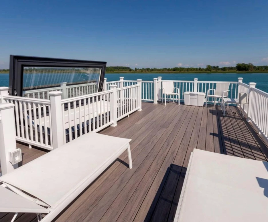 A spacious waterfront deck features white lounge chairs and railings, overlooking a calm blue lake under a clear blue sky.