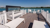 Terrasse moderne en bois avec des chaises longues blanches, offrant une vue sur l'eau calme et le ciel bleu. Des barrières blanches délimitent l'espace.