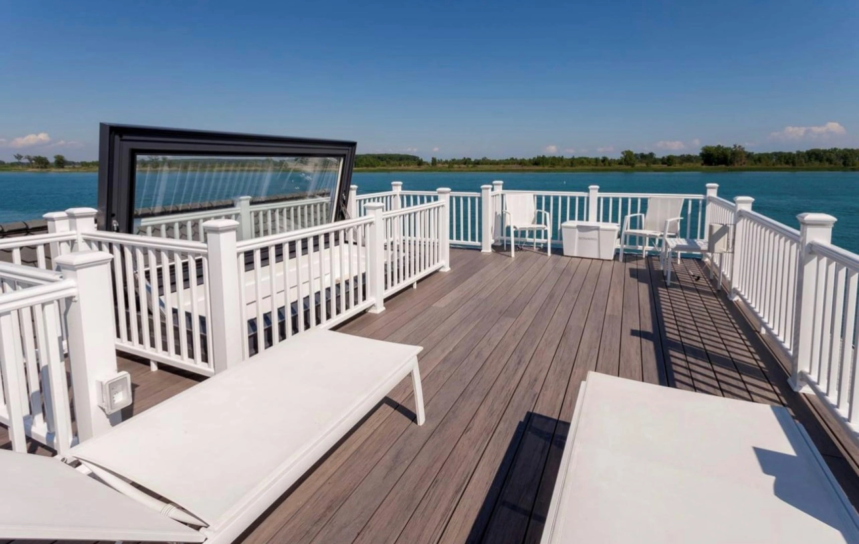 A spacious waterfront deck features white lounge chairs and railings, overlooking a calm blue lake under a clear blue sky.