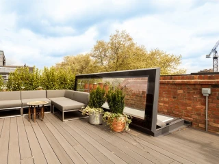 Terraza con muebles de exterior cómodos, incluyendo un sofá, una mesa y plantas en macetas. Al fondo, se aprecian árboles y un cielo despejado.