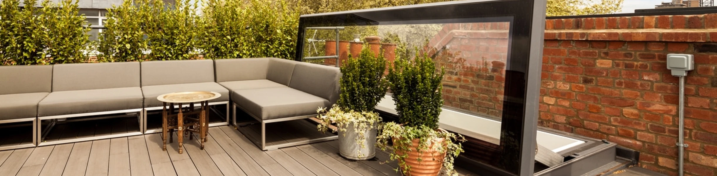 A stylish rooftop patio with gray seating, a round table, and potted plants. The space is surrounded by greenery and features a modern glass partition. Brick walls and a blue sky create a serene atmosphere.