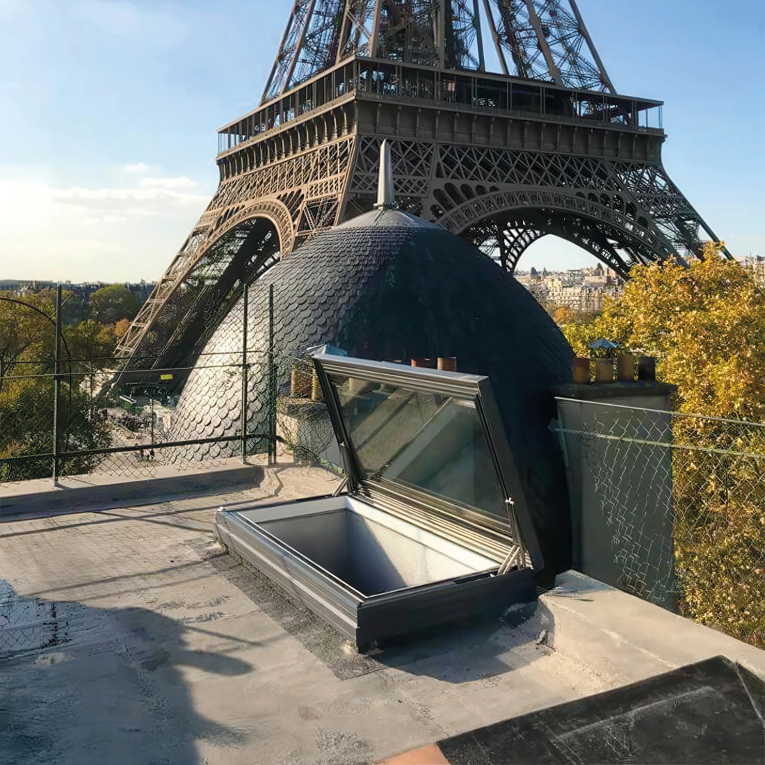 A rooftop view featuring the Eiffel Tower in the background, with a large dome and an open skylight in the foreground. Clear blue sky and autumn foliage are visible.