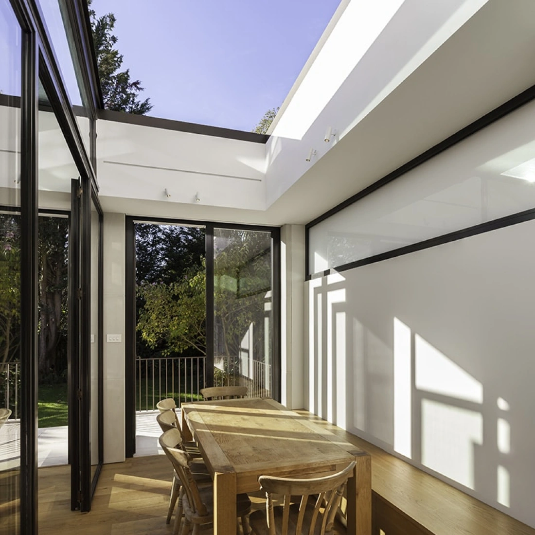 Comedor moderno con una mesa de madera clara y sillas de diseño sencillo. Las grandes ventanas permiten la entrada de luz natural y ofrecen vistas al jardín. Suelo de madera brillante y paredes blancas dan un aire minimalista al espacio.
