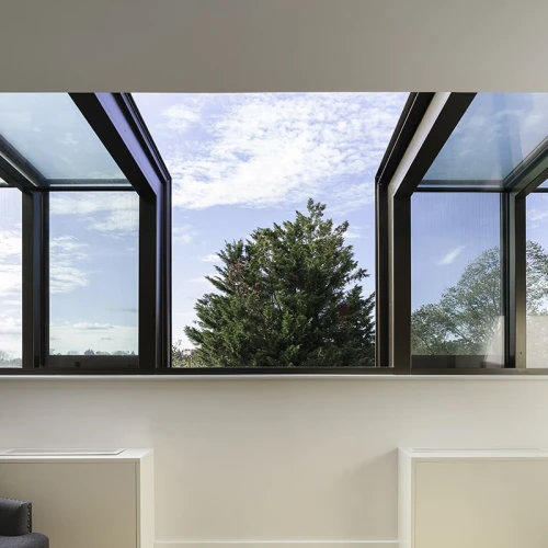 Des fenêtres ouvertes avec vue sur un arbre et un ciel partiellement nuageux. Un mur intérieur simple et deux radiateurs sont visibles en bas.