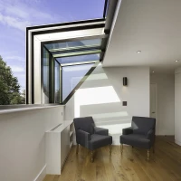 Deux chaises grises en tissu sont placées dans un coin d'une pièce moderne. Un grand vitrage permet une vue dégagée sur l'extérieur, sous un ciel bleu parsemé de nuages. Le sol en bois clair ajoute une touche chaleureuse à l'espace.