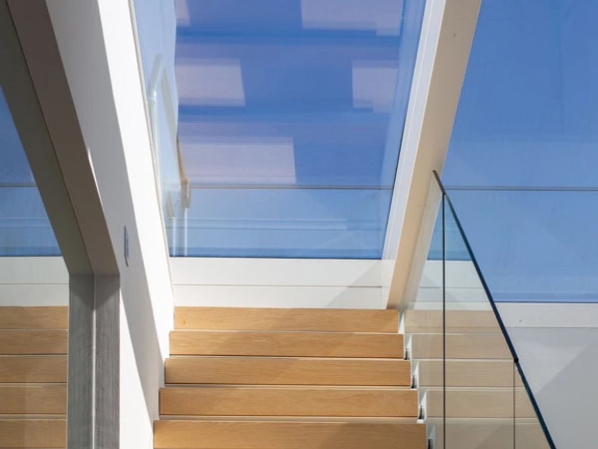 Light-filled staircase with wooden steps leading up to a skylight, featuring glass railings and modern architectural elements.