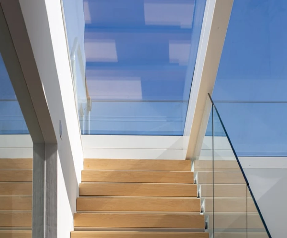 Light-filled staircase with wooden steps leading up to a skylight, featuring glass railings and modern architectural elements.