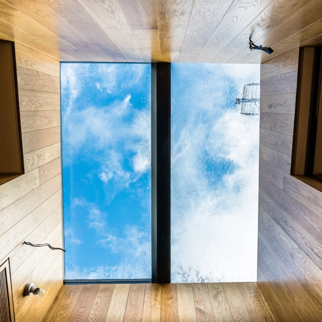 Vista desde el interior de una habitación con paredes de madera, mostrando una ventana rectangular en el techo que ofrece una vista del cielo nublado y algunos cables eléctricos.