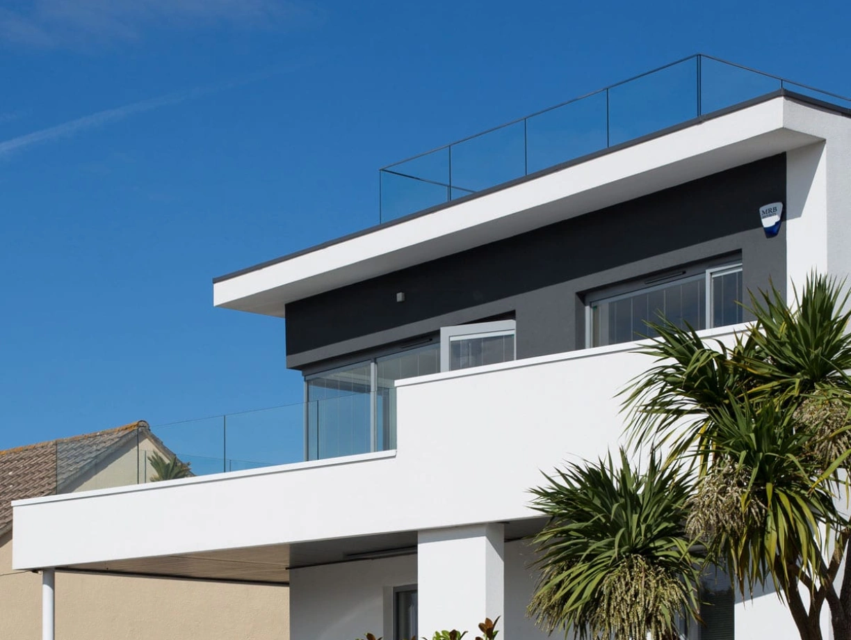 Casa moderna con un diseño minimalista, destacando una terraza con barandilla de vidrio. Jardines tropicales y un cielo despejado de fondo.